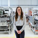 A picture of a smiling woman with engineering parts, presumably NDIR sensors, in the background on shelves