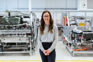 A picture of a smiling woman with engineering parts, presumably NDIR sensors, in the background on shelves