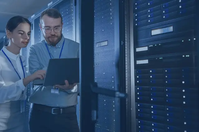 Two engineers discussing data on a laptop in a server room representing AI system monitoring