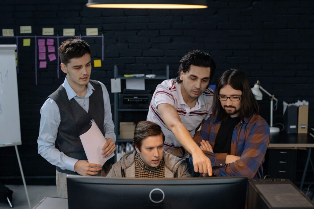 A group of four guys staring at a computer screen with a board of sticky notes behind them